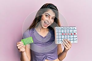 Young caucasian woman holding birth control pills and menstrual calendar smiling and laughing hard out loud because funny crazy