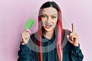 Young caucasian woman holding birth control pills smiling with an idea or question pointing finger with happy face, number one