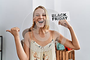 Young caucasian woman holding black friday banner at retail shop pointing thumb up to the side smiling happy with open mouth