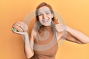 Young caucasian woman holding brain smiling happy and positive, thumb up doing excellent and approval sign