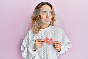 Young caucasian woman holding broken heart paper shape smiling looking to the side and staring away thinking