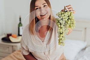 Young Caucasian Woman Holding Bunch of Grapes in Hand While Sitting in Hotel Room, Tray of Fruit and Bottle of Wine