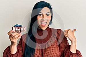 Young caucasian woman holding cake slice pointing thumb up to the side smiling happy with open mouth