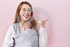 Young caucasian woman holding and carrying baby on a sling pointing thumb up to the side smiling happy with open mouth