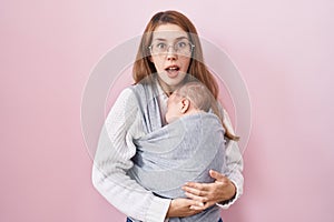 Young caucasian woman holding and carrying baby on a sling scared and amazed with open mouth for surprise, disbelief face