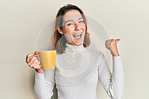 Young caucasian woman holding coffee pointing thumb up to the side smiling happy with open mouth