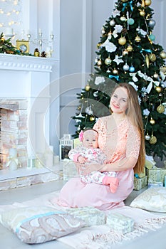 Young caucasian woman holding daughter in arms near decorated fireplace and Christmas tree, wearing pink dress.