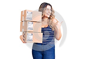 Young caucasian woman holding delivery package pointing thumb up to the side smiling happy with open mouth