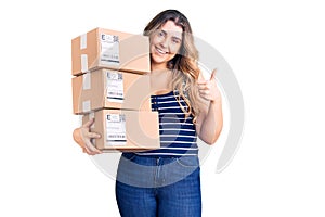 Young caucasian woman holding delivery package smiling happy and positive, thumb up doing excellent and approval sign