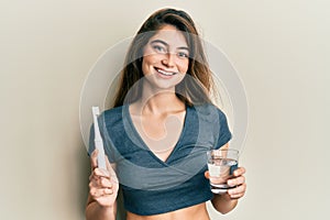 Young caucasian woman holding electric toothbrush and water glass smiling with a happy and cool smile on face