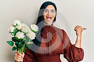 Young caucasian woman holding flowers pointing thumb up to the side smiling happy with open mouth