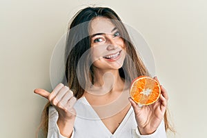 Young caucasian woman holding fresh slice of orange pointing thumb up to the side smiling happy with open mouth