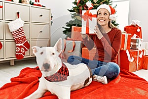 Young caucasian woman holding gift sitting with dog by christmas tree at home