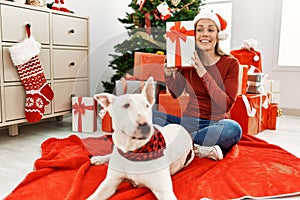 Young caucasian woman holding gift sitting with dog by christmas tree at home