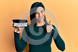 Young caucasian woman holding glasses smiling happy and positive, thumb up doing excellent and approval sign