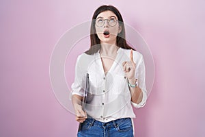 Young caucasian woman holding laptop amazed and surprised looking up and pointing with fingers and raised arms