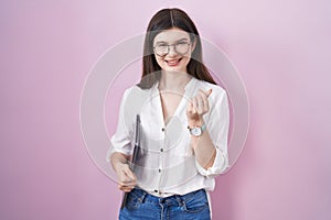 Young caucasian woman holding laptop doing money gesture with hands, asking for salary payment, millionaire business