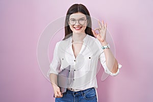 Young caucasian woman holding laptop smiling positive doing ok sign with hand and fingers