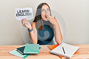 Young caucasian woman holding learn english paper while studying covering mouth with hand, shocked and afraid for mistake