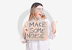 Young caucasian woman holding make some noise banner pointing thumb up to the side smiling happy with open mouth