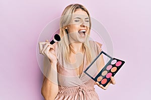 Young caucasian woman holding makeup brush and blush palette angry and mad screaming frustrated and furious, shouting with anger