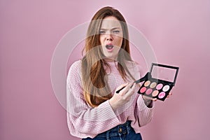 Young caucasian woman holding makeup brush and blush palette angry and mad screaming frustrated and furious, shouting with anger