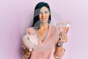 Young caucasian woman holding norwegian krona banknotes and piggy bank clueless and confused expression