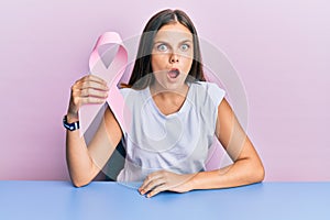 Young caucasian woman holding pink cancer ribbon scared and amazed with open mouth for surprise, disbelief face