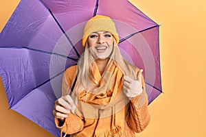 Young caucasian woman holding purple umbrella smiling happy and positive, thumb up doing excellent and approval sign