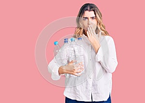 Young caucasian woman holding recycling plastic bottles covering mouth with hand, shocked and afraid for mistake
