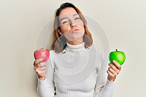 Young caucasian woman holding red and green apple looking at the camera blowing a kiss being lovely and sexy