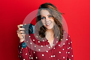 Young caucasian woman holding reflex camera looking positive and happy standing and smiling with a confident smile showing teeth