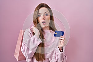 Young caucasian woman holding shopping bag and credit card afraid and shocked with surprise and amazed expression, fear and