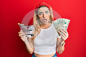 Young caucasian woman holding small supermarket shopping cart and dollars in shock face, looking skeptical and sarcastic,