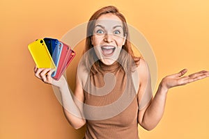 Young caucasian woman holding smartphone case celebrating achievement with happy smile and winner expression with raised hand