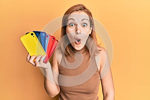 Young caucasian woman holding smartphone case scared and amazed with open mouth for surprise, disbelief face