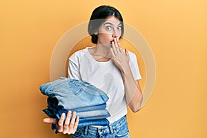 Young caucasian woman holding stack of folded jeans covering mouth with hand, shocked and afraid for mistake