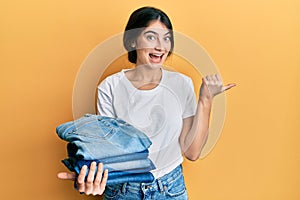 Young caucasian woman holding stack of folded jeans pointing thumb up to the side smiling happy with open mouth