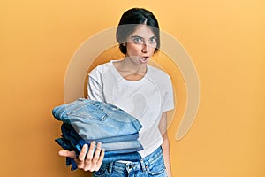 Young caucasian woman holding stack of folded jeans scared and amazed with open mouth for surprise, disbelief face