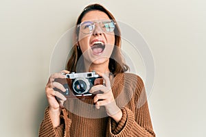 Young caucasian woman holding vintage camera angry and mad screaming frustrated and furious, shouting with anger looking up