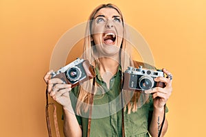 Young caucasian woman holding vintage camera angry and mad screaming frustrated and furious, shouting with anger looking up