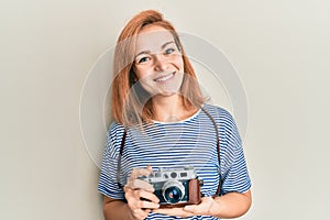 Young caucasian woman holding vintage camera looking positive and happy standing and smiling with a confident smile showing teeth
