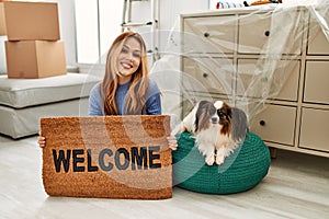 Young caucasian woman holding welcome doormat sitting on floor with dog at new home