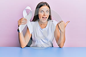 Young caucasian woman holding white ribbon pointing thumb up to the side smiling happy with open mouth