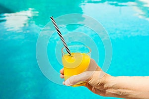 Young Caucasian Woman Holds Glass of Freshly Pressed Orange Juice with Black and White Striped Straw by Swimming Pool Summer