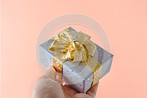 Young Caucasian woman holds in hand gift box wrapped in silver paper with golden ribbon bow on pink background. Christmas