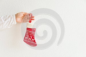 Young caucasian woman holds in hand knitted red Christmas gift stocking on white wall background. Family corporate presents