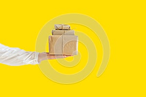Young Caucasian woman holds hand stack of boxes wrapped in brown paper tied with twine on yellow background. Contactless delivery