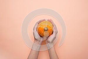 Young Caucasian Woman Holds in Hands Small Round Heirloom Orange Pumpkin on Peachy Pink Background. Fall Autumn