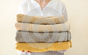 Young caucasian woman holds in hands stack of clean folded terry towels. White wall background. Laundry spa wellness cleanliness